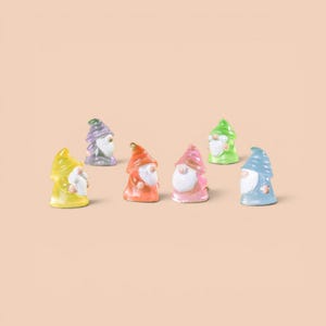 May include: A collection of seven colorful garden gnomes. Each gnome has a pointed hat and a long white beard. The gnomes are in shades of yellow, purple, orange, pink, green, and blue. They are arranged on a solid peach-colored background.
