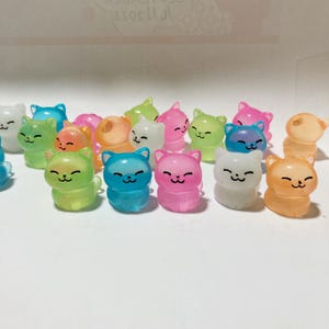 May include: A collection of small, colourful, translucent cat figurines. The cats are in various poses and have a cute, cartoonish design. They are made of a clear plastic material and glow in the dark.