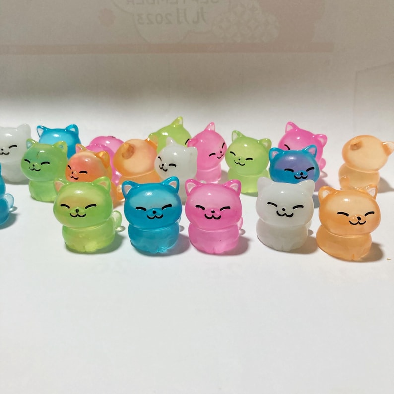 May include: A collection of small, colourful, translucent cat figurines. The cats are in various poses and have a cute, cartoonish design. They are made of a clear plastic material and glow in the dark.