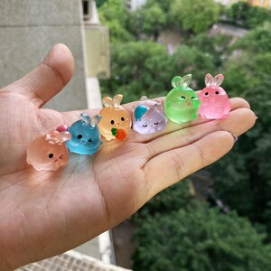 Op de afbeelding: Zes kleine, kleurrijke, doorzichtige plastic konijnenfiguren. De konijnen staan in verschillende poses en hebben verschillende gekleurde accenten. De konijnen zijn in een rij op een hand gerangschikt.