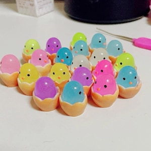 May include: A collection of colorful, miniature chicks in eggshells. The chicks are made of clear plastic and have painted details. The eggshells are made of a white material and have a textured surface.