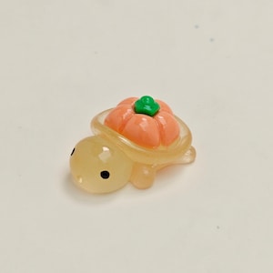 May include: A small, translucent turtle figurine with a pumpkin shell. The turtle is light yellow with black eyes. The pumpkin is orange with a green stem. The figurine is likely made of resin or a similar material.