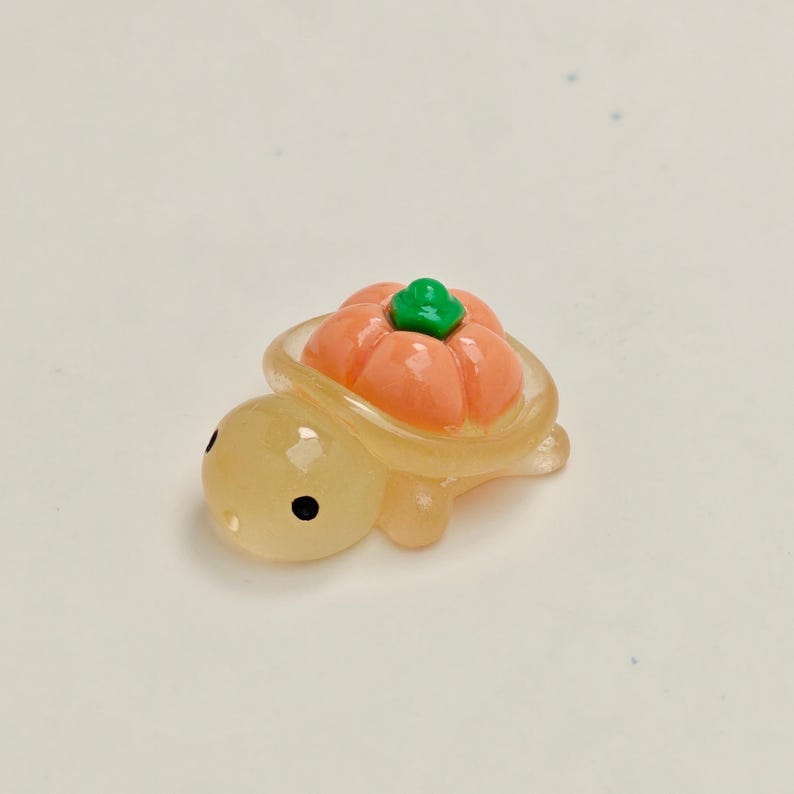 May include: A small, translucent turtle figurine with a pumpkin shell. The turtle is light yellow with black eyes. The pumpkin is orange with a green stem. The figurine is likely made of resin or a similar material.