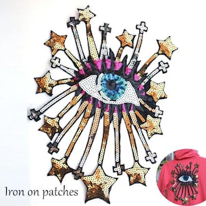 May include: Iron-on patch featuring a large, colorful eye with sequins and gold star accents. The eye is surrounded by a burst of gold stars and sequins. The patch is iron-on and can be used to decorate clothing, bags, and other items. "Iron on patches" is written below the patch.