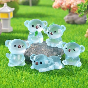 Könnte beinhalten: Sechs durchscheinende blaue Koala-Bärenfiguren auf einer grasbewachsenen Oberfläche. Die Koalas haben braune Nasen und schwarze Augen. Einige stehen, einige sitzen und einer liegt. Jeder Koala hat ein kleines grünes Blatt auf dem Kopf.