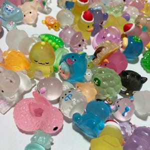 Könnte beinhalten: Eine Sammlung von bunten, durchsichtigen und detaillierten Miniaturfiguren. Die Figuren sind aus klarem Kunststoff gefertigt und zeigen verschiedene Tier- und Charakterdesigns. Einige der Figuren haben Glitzer oder andere dekorative Elemente.
