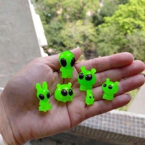 May include: Five small, translucent neon green figurines with smooth surfaces and large, solid black oval eyes. The collection includes a standing alien, a rabbit-like creature, a round blob shape, a larger alien, and a tiny alien figure.