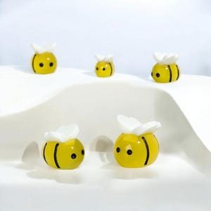 Puede incluir: Cinco pequeñas figuras de abejas amarillas con rayas negras y alas blancas están dispuestas sobre una superficie blanca. Las abejas tienen ojos negros y están en varias posiciones. Las figuras son probablemente artículos decorativos.