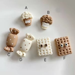 Puede incluir: Colección de seis colgantes en miniatura con temática de comida. Los colgantes incluyen dos cupcakes, dos caramelos envueltos con lunares y dos galletas saladas con caras de dibujos animados. Los colores son principalmente crema, marrón y beige.