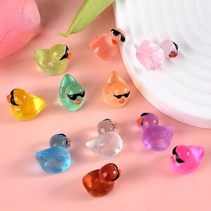 May include: A collection of 12 colorful rubber duck figurines with sunglasses. The ducks are in various colors, including yellow, green, blue, pink, orange, and clear. They are all wearing black sunglasses.
