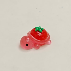 May include: A small, pink turtle figurine with a red tomato shell and green leaves on top. The turtle has a black eye and is made of a glossy material. This whimsical figurine is a charming decorative item.