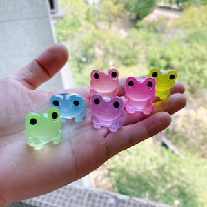 以下が含まれることがあります： 透明なプラスチック製の小さなカラフルなカエルの置物6個。カエルは緑、青、ピンク、黄色の様々な色合いです。
