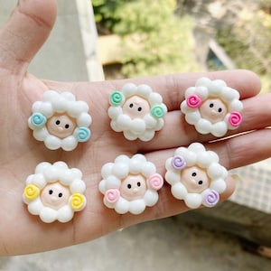 May include: Six white sheep-shaped resin embellishments with different colored flowers. Each sheep has a pink face and black eyes. The flowers are in shades of pink, purple, green, and yellow.