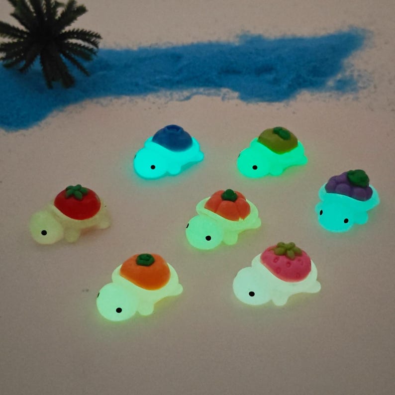 May include: Seven glowing turtle figurines with fruit-shaped shells are arranged on a light background. The turtles have various fruit designs, including a tomato, orange, strawberry, and grapes. The turtles glow with a soft, inviting light.
