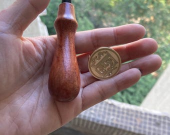 Puede incluir: Un mango de madera para un sello de cera con un sello de cera de metal dorado con un diseño decorativo.