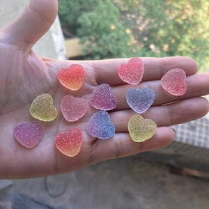 Op de afbeelding: Een verzameling hartvormige snoepjes in verschillende pastelkleuren, waaronder roze, blauw, geel en paars. De snoepjes zijn op een hand gelegd, waardoor hun levendige kleuren en suikerachtige textuur worden getoond.