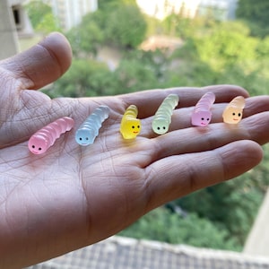 May include: A hand holding six small, colorful, gummy worm-shaped toys. The worms are in a rainbow of colors, including pink, blue, green, yellow, and orange. Each worm has a smiling face.