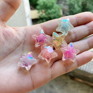 Pode incluir: Uma coleção de contas iridescentes em forma de estrela em vários tons de rosa, amarelo e transparente. As contas têm uma qualidade brilhante e translúcida e são seguradas em uma mão.