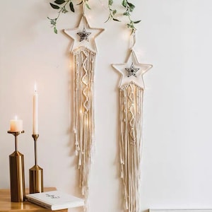 Op de afbeelding: Twee macramé ster wanddecoraties met lichtsnoeren. De sterren zijn wit met een zwart sterontwerp in het midden. De decoraties zijn gemaakt van natuurlijke katoenen koord en hangen aan een witte muur.