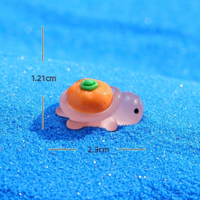 May include: A miniature turtle figurine, 2.3 cm long and 1.21 cm tall, with a translucent lavender body and an orange shell topped with a green accent. The figurine is set against a blue, textured background.