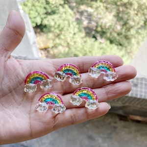 Pode incluir: Cinco enfeites em forma de arco-íris com detalhes de glitter. Cada arco-íris apresenta cores rosa, amarelo, azul e verde, com uma nuvem na parte inferior. Os enfeites são pequenos e parecem ser feitos de resina ou plástico.