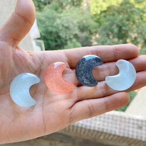 Puede incluir: Cuatro amuletos de resina en forma de media luna con purpurina incrustada. Los amuletos son de color azul claro, rosa, negro y blanco.