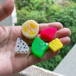 May include: Five colorful fruit-shaped silicone molds, including a yellow pineapple, a green kiwi, a pink watermelon, a white and black strawberry, and a yellow banana.