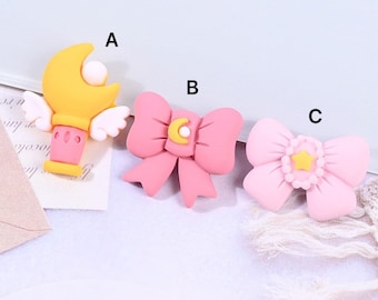 May include: Three pink and yellow resin charms with a moon and star design. The charms are shaped like a wand, a bow, and a bow with a star in the center.