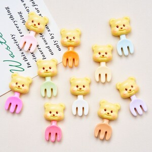 May include: Ten colorful plastic forks with bear faces. The forks are yellow with different colored handles: pink, orange, light green, white, light blue, purple, and a darker shade of green.