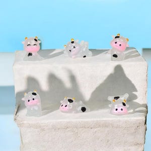 Pode incluir: Seis miniaturas de vacas em várias poses. As vacas são translúcidas com rostos rosa, manchas pretas e pequenos chifres amarelos. Elas estão dispostas em uma superfície branca texturizada contra um fundo azul claro.