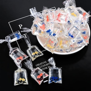 May include: Assortment of small clear plastic bags filled with colorful goldfish-shaped charms. The bags are tied with a ribbon and are perfect for adding a whimsical touch to crafts, jewelry, or party favors.