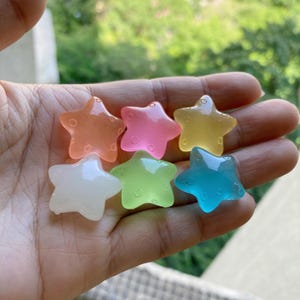 May include: Six colorful star-shaped resin charms in shades of orange, pink, yellow, white, green, and blue. Each star has a smooth, translucent appearance and is held in a hand against a blurred green background.