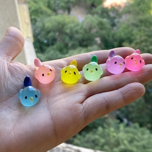 Könnte beinhalten: Sechs kleine Glasfiguren in Form von Küken in verschiedenen Pastellfarben, darunter Blau, Rosa, Gelb und Grün. Die Figuren sind in einer Reihe auf einer Hand angeordnet.