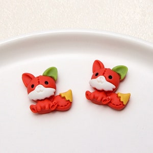 Puede incluir: Dos encantos de arcilla polimérica en forma de zorro rojo con orejas verdes y colas amarillas. Los zorros tienen caras blancas con ojos negros y detalles cosidos.