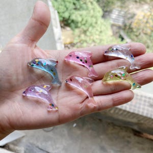 Puede incluir: Seis pequeñas figuritas de delfines hechas de resina transparente con purpurina incrustada en el interior. Los delfines son de varios colores, incluyendo rosa, azul, verde y transparente.