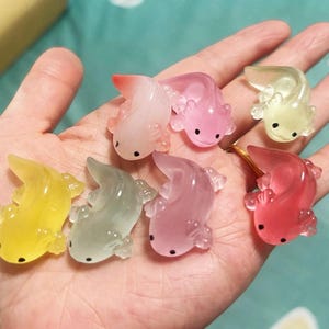以下が含まれることがあります： ピンク、黄色、緑、赤の色合いのカラフルなウーパールーパーのフィギュアのコレクション。各ウーパールーパーは、目に小さな黒い点があります。フィギュアは半透明で、樹脂またはガラスでできているようです。