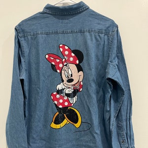 Könnte beinhalten: Ein blaues Jeanshemd mit einem Vintage-Minnie-Mouse-Motiv auf der Rückseite. Das Motiv zeigt Minnie Mouse in einem roten, gepunkteten Kleid und einer roten Schleife mit weißen Punkten.