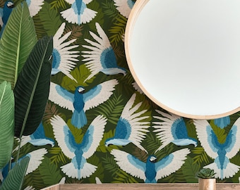 May include: Wallpaper featuring a repeating pattern of blue and white macaws flying amidst dark green tropical foliage. A round mirror with a light wood frame hangs above a white cabinet with a small succulent plant. The text Paisley & Dot is visible.