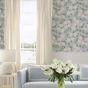 May include: A light blue living room with cream curtains framing a window overlooking a lake. A light blue sofa with white trim is in front of the window. A white lamp and a vase of white flowers are on a table.