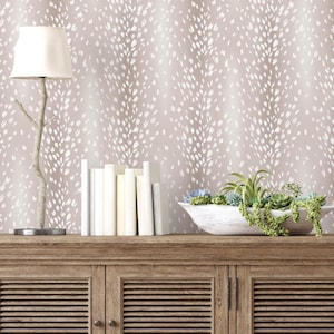 May include: A light brown wallpaper with a white abstract pattern. The wallpaper is displayed on a wall behind a wooden cabinet with two doors. The cabinet has a white lamp with a white shade on the left side and a white bowl with green plants on the right side.