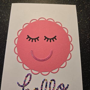 May include: A white greeting card with a pink scalloped circle featuring closed black eyelashes and a glittery smile. The word "hello" is written in colorful glittery script below the circle.