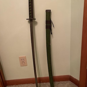 May include: A katana sword with a dark blade and a green and brown wrapped handle. The scabbard is green with brown accents. The sword is displayed vertically against a wall.