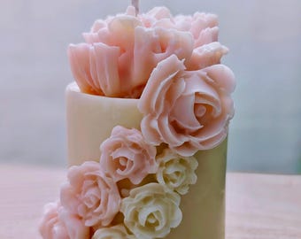 Può includere: Una candela cilindrica con un design floreale. La candela è di colore giallo pallido, con un gruppo di petali di garofano rosa sulla parte superiore. Rose rosa e bianche scendono a cascata sul lato e uno stoppino bianco si estende dalla parte superiore.
