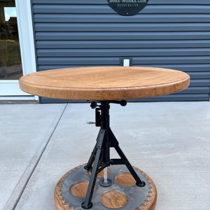 May include: A round wooden table with a black metal base and a gear-shaped base. The table is made of oak wood and has a natural finish. The base is adjustable in height. The table is 24 inches in diameter and 30 inches tall.