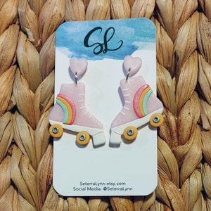 May include: A pair of pink roller skate earrings with rainbow accents and yellow wheels. The earrings are on a white card with the text "Seterralynn.etsy.com" and "Social Media: @Seterralynn".