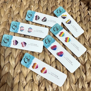 May include: A set of eight pairs of heart-shaped earrings with different pride flag designs. The earrings are displayed on small white cards with the text "Seterralynn.etsy.com" and "@Seterralynn".