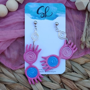 May include: Pink and blue glitter earrings shaped like a pair of glasses with a spiral design. The earrings are hanging from silver hooks with a spiral charm.