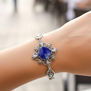 May include: Silver bracelet featuring a large, square, deep blue gemstone at its center, surrounded by an ornate, openwork design. The bracelet is worn on a wrist, with a blurred outdoor background.