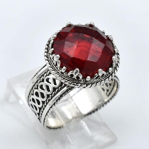 May include: A silver ring with a large, faceted, deep red gemstone. The ring has an ornate, filigree design on the band and around the gemstone. The gemstone is approximately 0.75 inches in diameter.