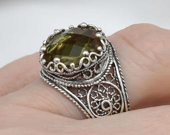 May include: A silver ring featuring an olive-green gemstone. The ring has an ornate filigree design around the stone and on the band. The gemstone is oval-shaped and set in a detailed, crown-like setting. The ring is on a finger.